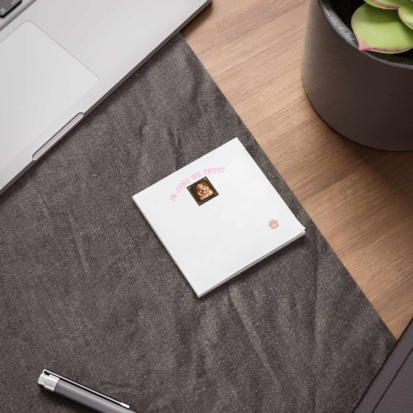 Small white sticky note pad on a textured gray surface with a laptop and flowering plant in the background.