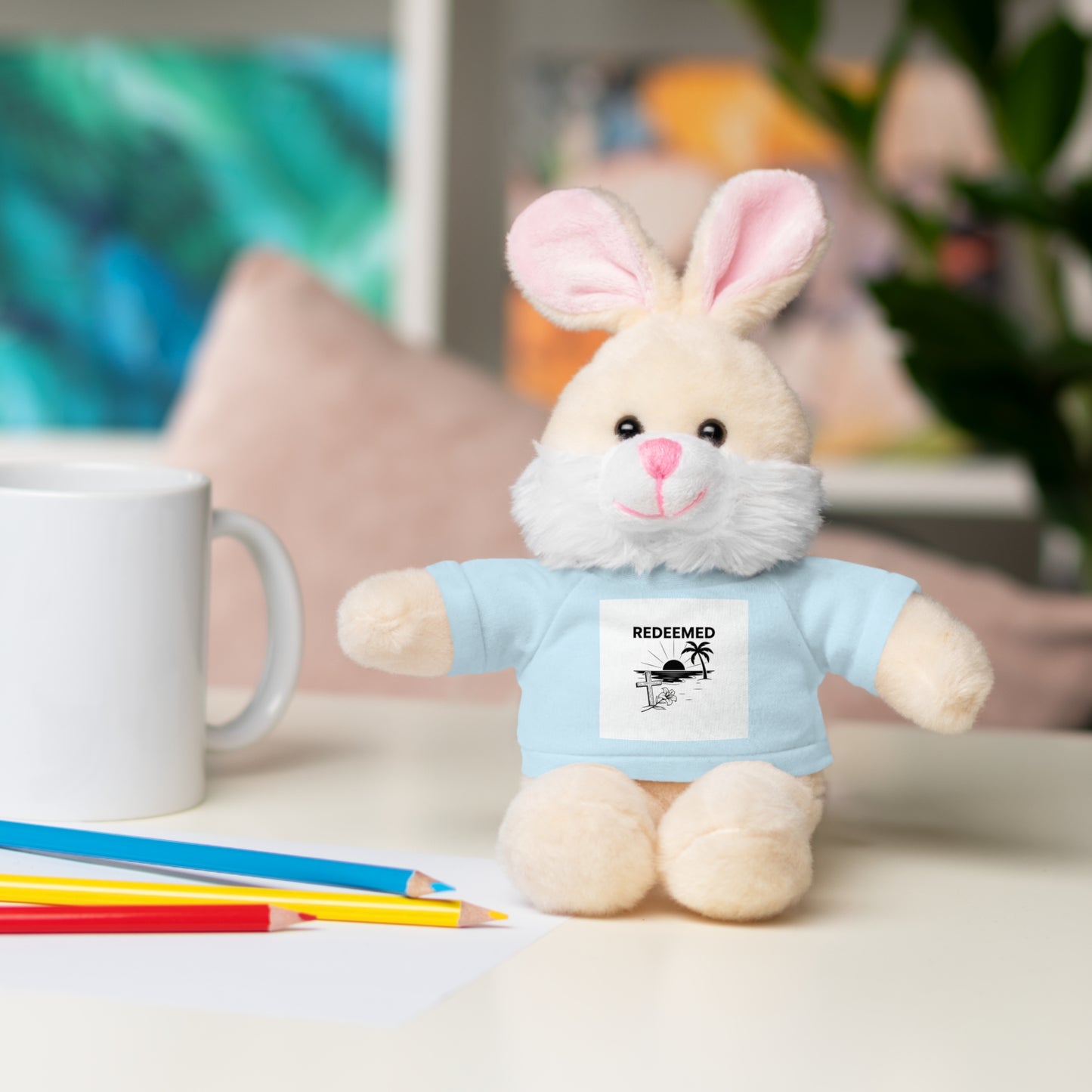 Plush bunny toy wearing a blue shirt with text, sitting on a table with coloring pencils and a mug.