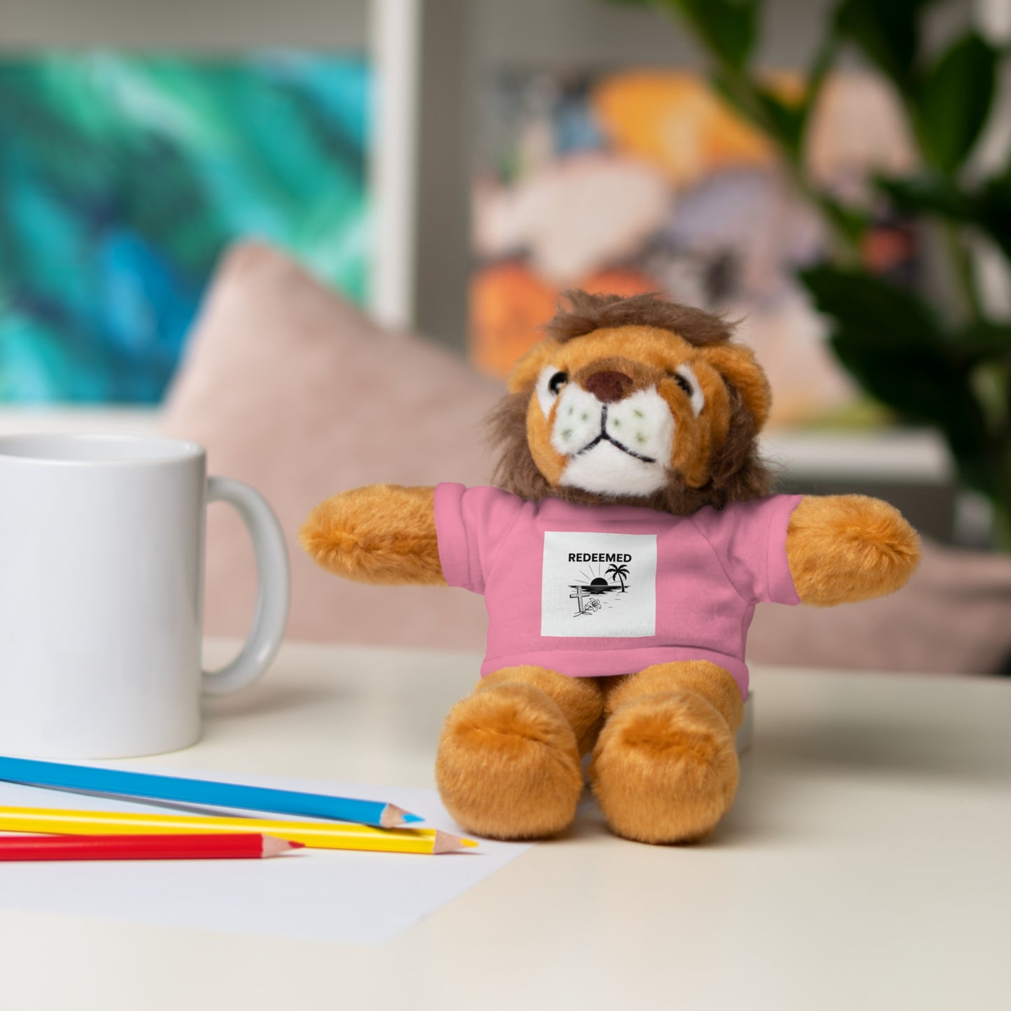 Plush lion toy wearing a pink shirt with a label, sitting on a surface with a mug and pencils in the background.