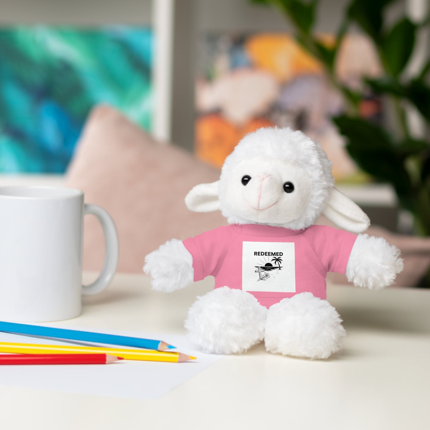 White plush lamb toy wearing a pink shirt with a label, sitting on a table with a mug and pencils.