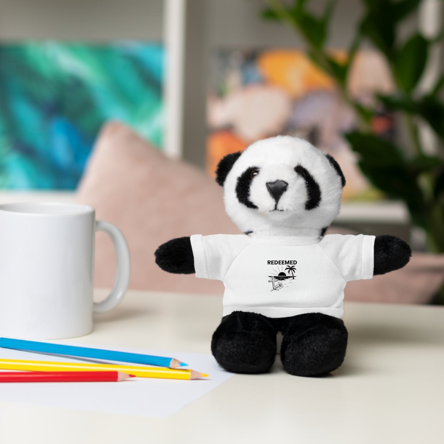 Plush panda toy wearing a white t-shirt with text, placed on a surface with colored pencils and a mug.