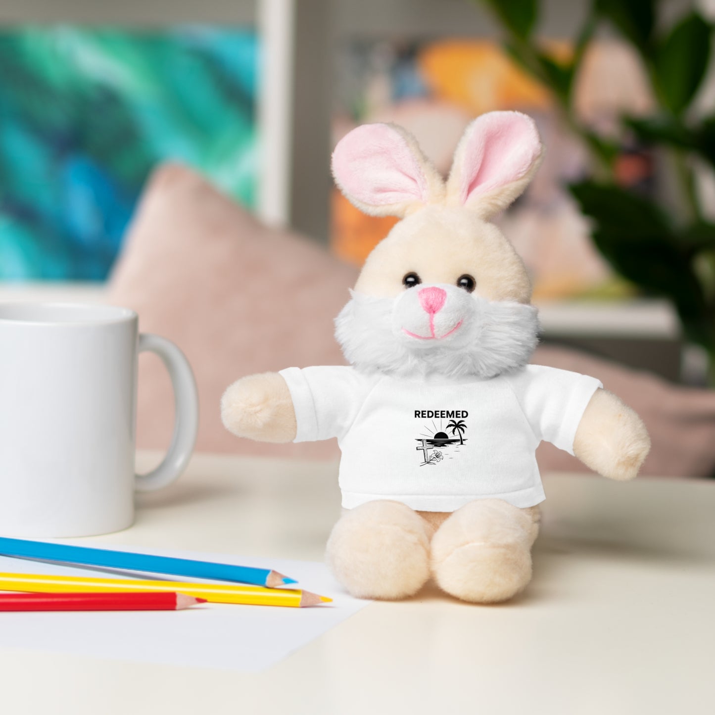 Plush toy rabbit wearing a white t-shirt with text, sitting on a surface with coloring pencils and a mug.