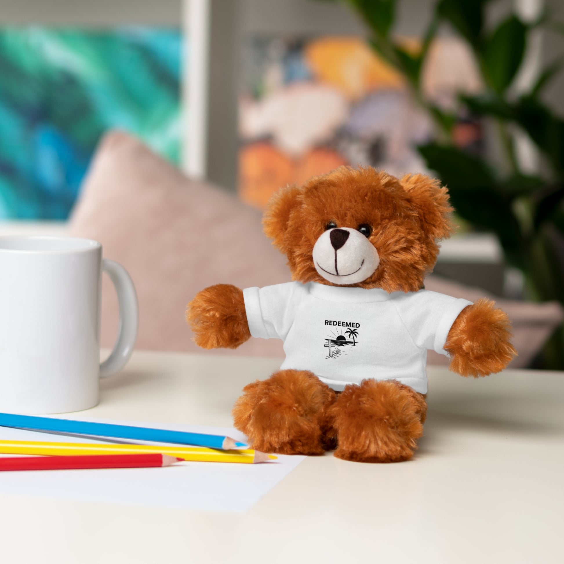 Brown teddy bear wearing a white t-shirt with text, sitting on a table with stationery items.