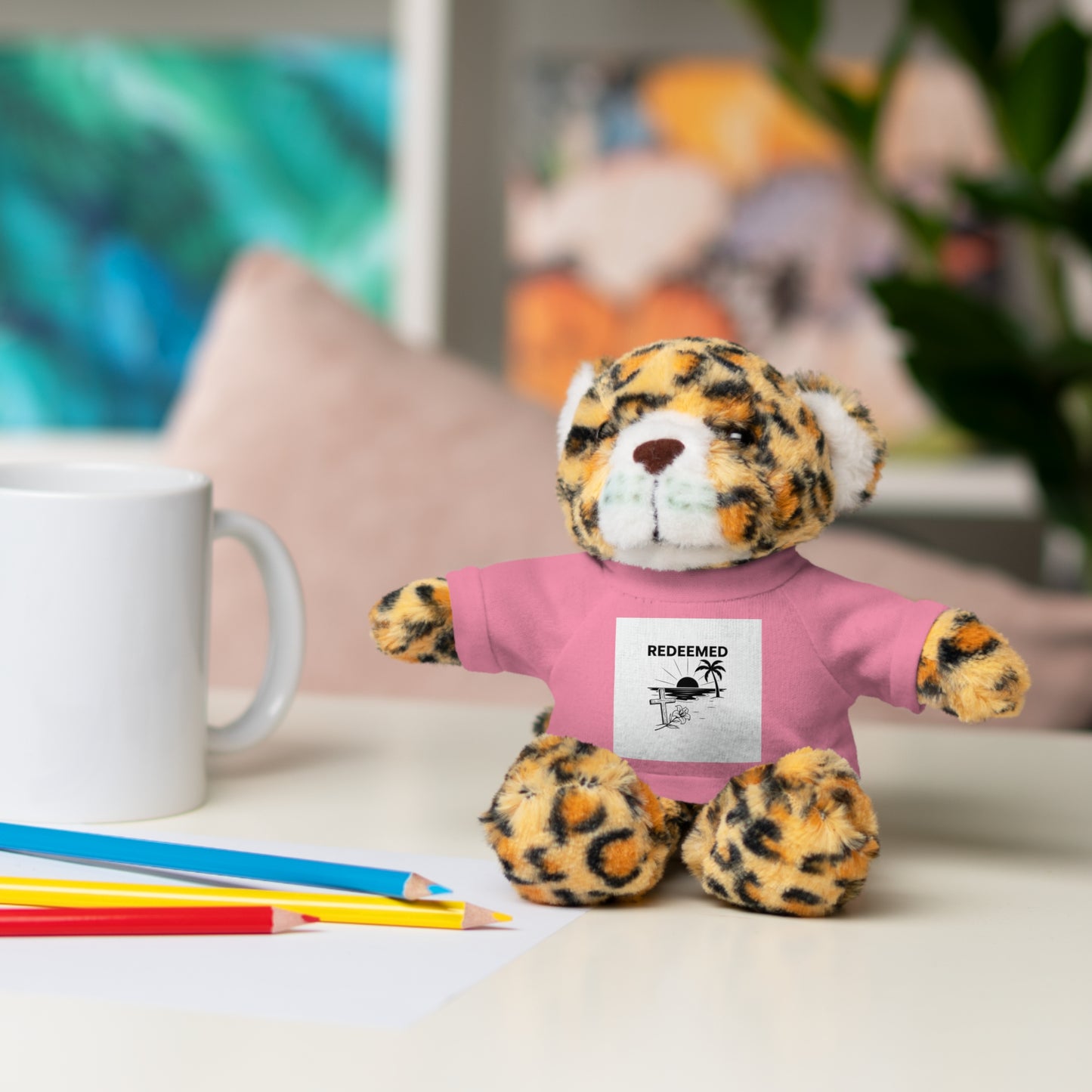 Plush toy leopard wearing a pink shirt with text on a desk with stationery items.