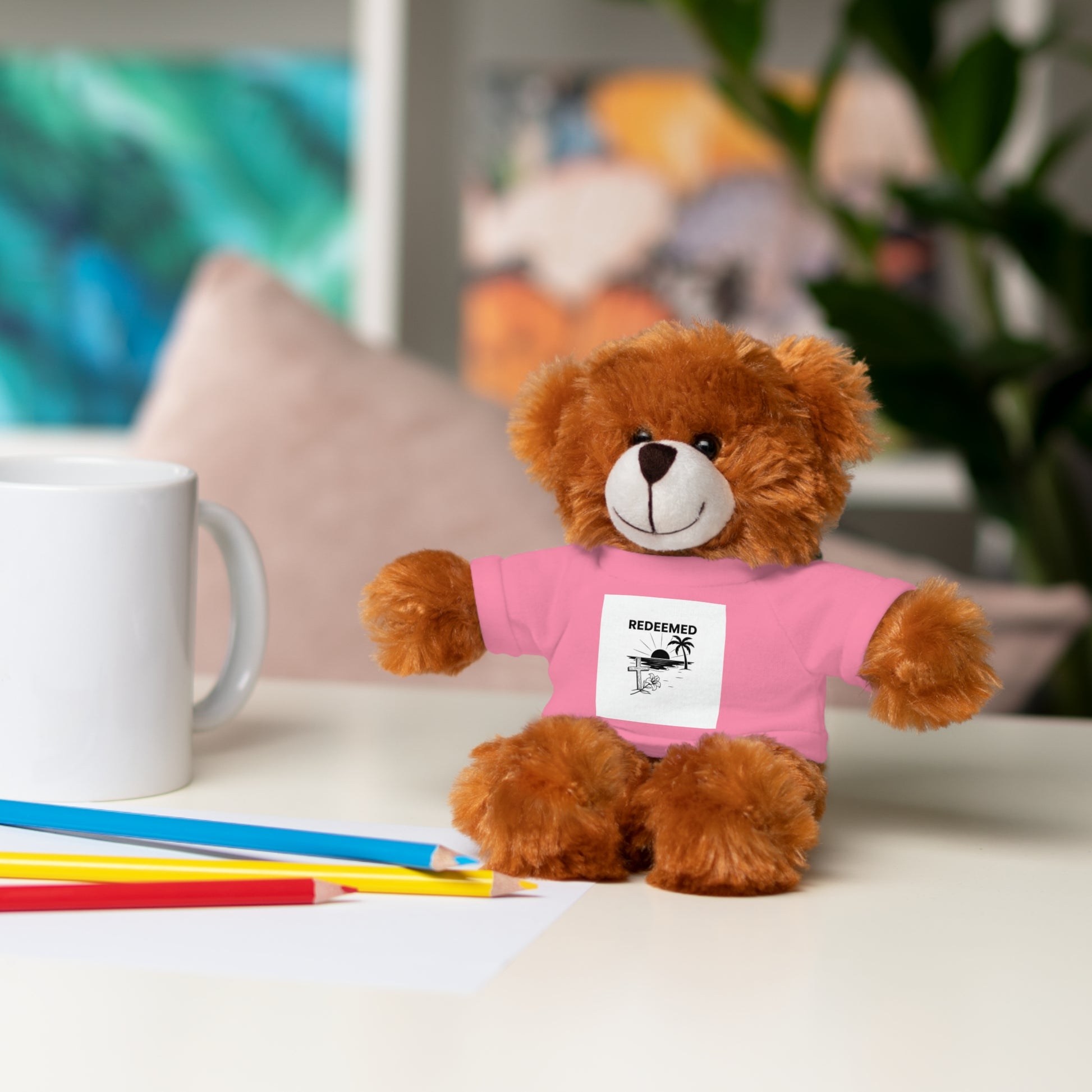 Brown teddy bear wearing a pink shirt with text, sitting on a table with a mug and pencils.