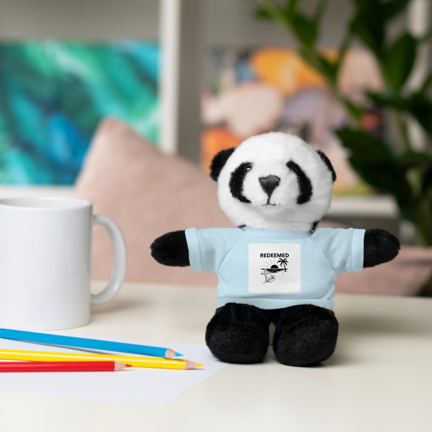 Plush panda toy wearing a blue shirt with text, sitting on a table with coloring pencils and a mug.