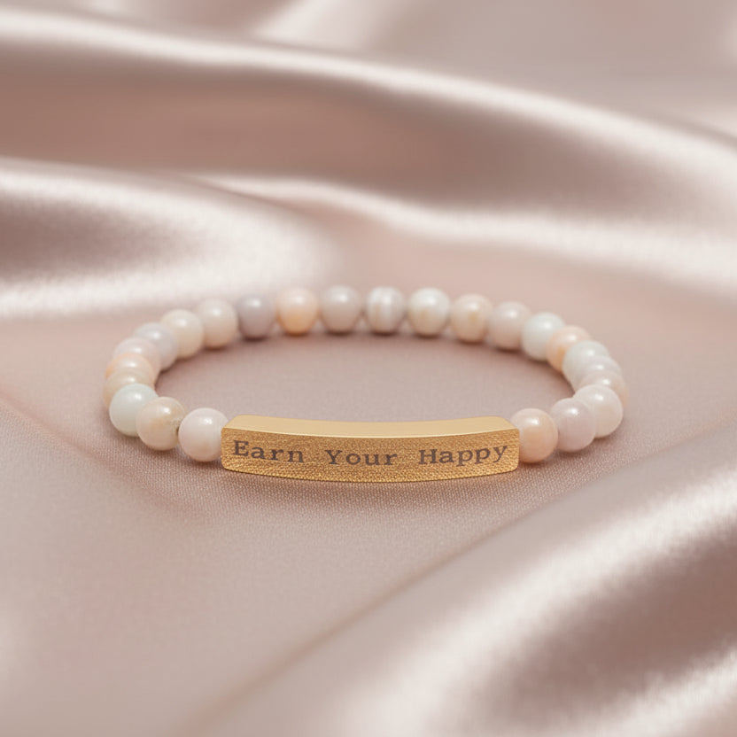 Natural Stone stretch bracelet with a gold bar message "Earn Your Happy on a blush pink satin surface