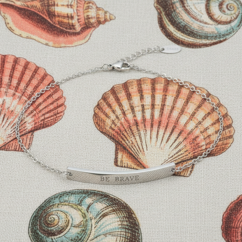 Silver anklet with 'BE BRAVE' engraving on a background of illustrated shells.
