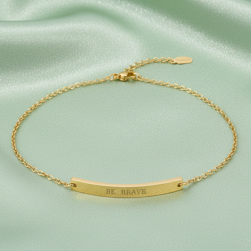 Gold anklet with 'BE BRAVE' engraving on a light green satin background

