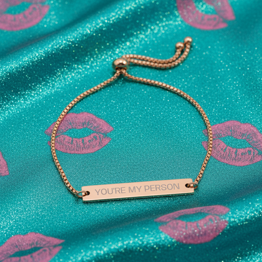 Gold bracelet with 'YOU'RE MY PERSON' engraving on a turquoise fabric background with pink lip prints.