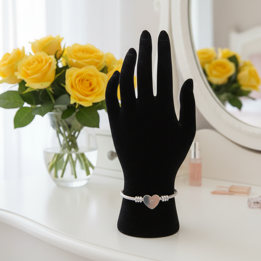 Black jewelry display mannequin hand with a silver heart-shaped bracelet with Initial "C" yellow roses in the background