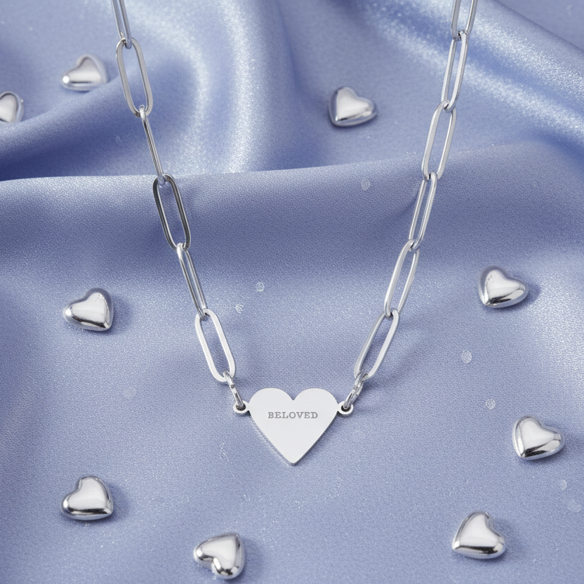 Silver necklace with a heart-shaped pendant labeled 'Beloved' on a blue satin background with heart-shaped objects.