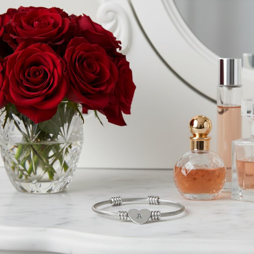 Silver bracelet with heart charm, engraved with the initial "A" and  red roses in a vase, and perfume bottles on a marble surface.