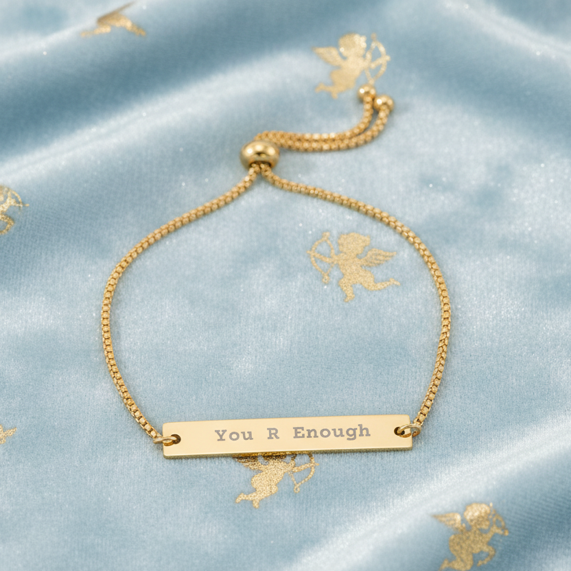 Gold bracelet with 'You R Enough' bar on a light blue satin background