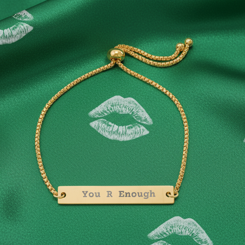 Gold bracelet with 'You R Enough' bar on a green fabric background with lip prints.