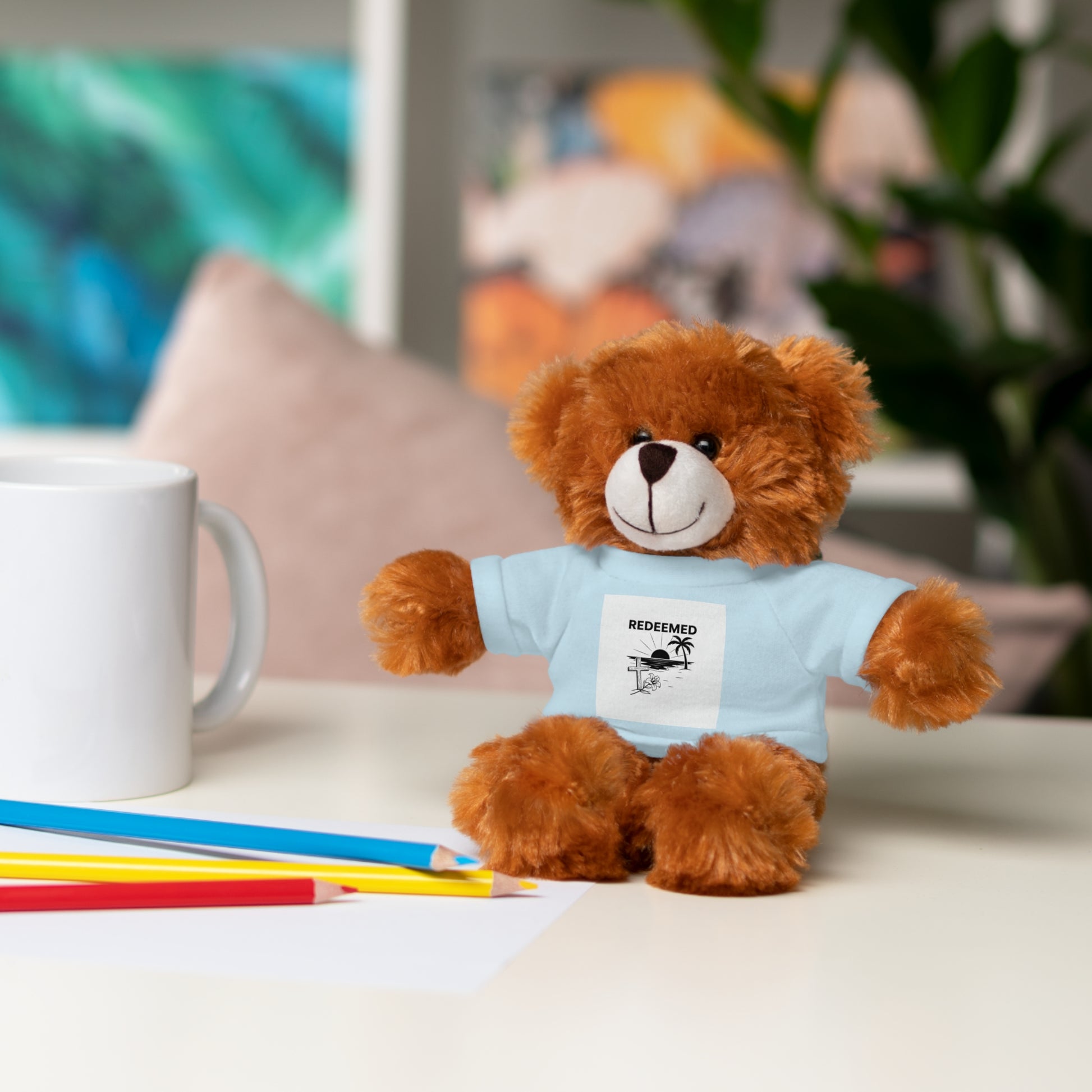 Brown teddy bear wearing a blue t-shirt with text, sitting on a table with stationery items.