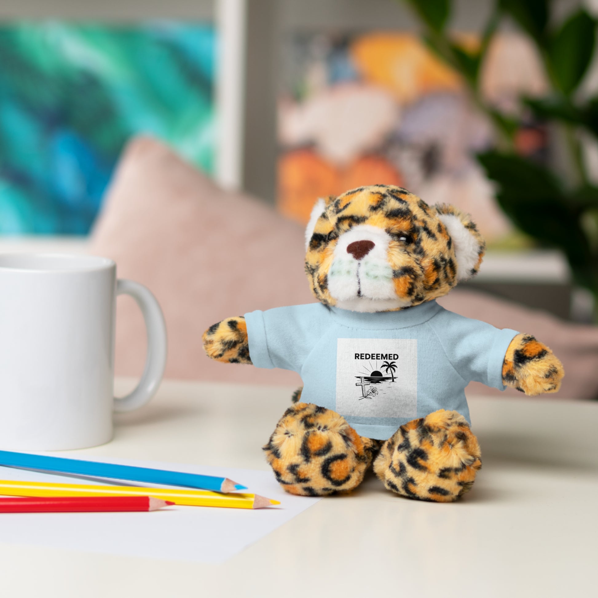 Leopard print teddy bear wearing a blue shirt with text, sitting on a desk with stationery items