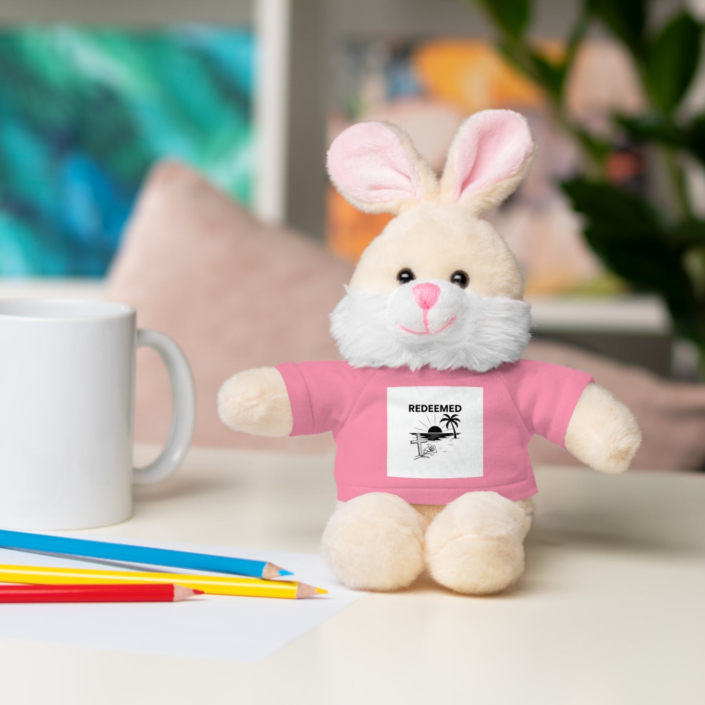 Plush toy rabbit wearing a pink shirt with a label on a table with colored pencils and a mug.