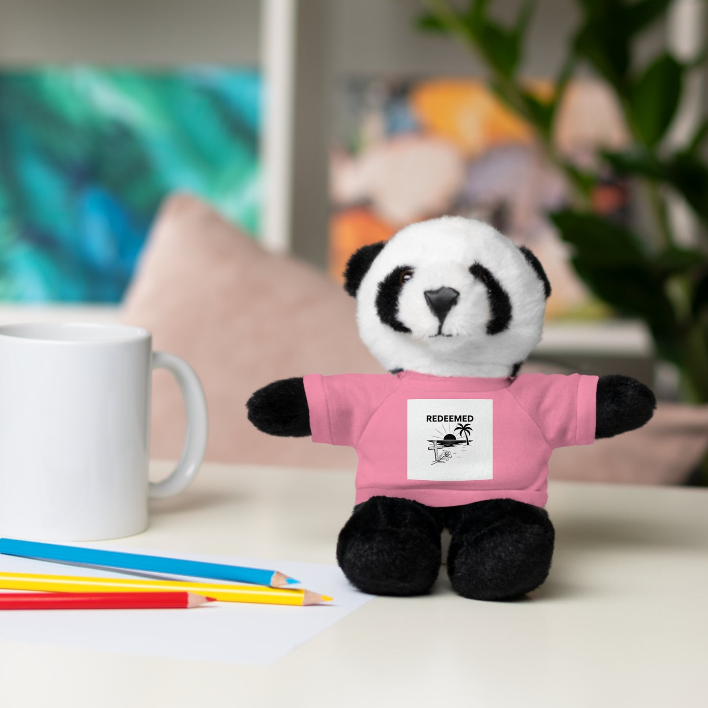 Plush panda toy wearing a pink shirt with a graphic on a table with coloring supplies.