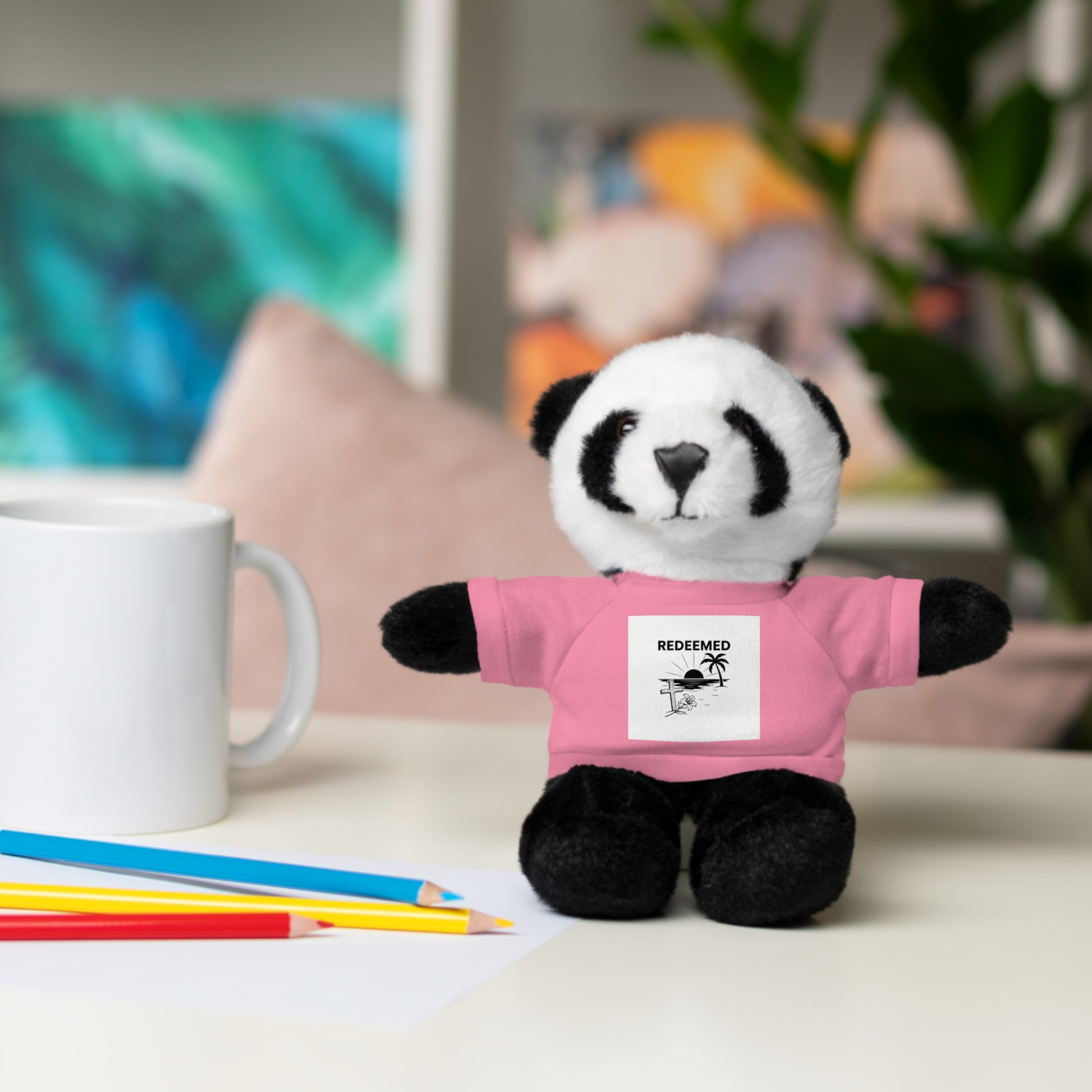 Plush panda toy wearing a pink shirt with a graphic on a table with coloring supplies.