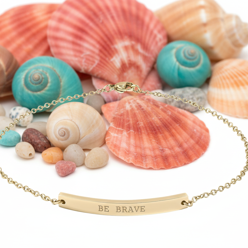Gold anklet with 'BE BRAVE' engraving on a background of seashells and pebbles.
