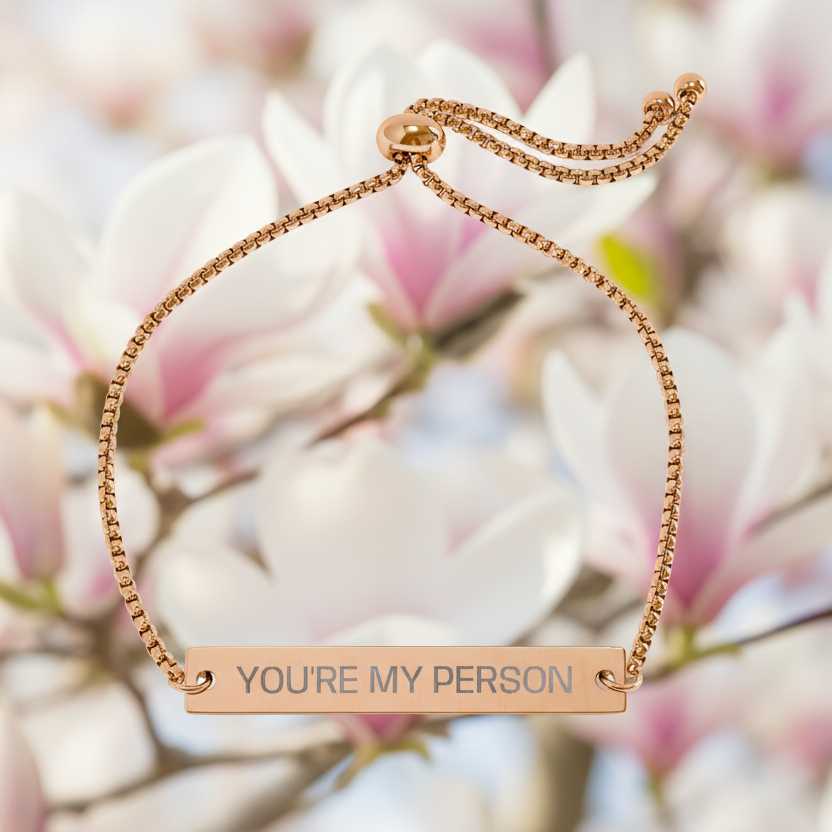 Rose gold bracelet with 'YOU'RE MY PERSON' engraving against a blurred floral background