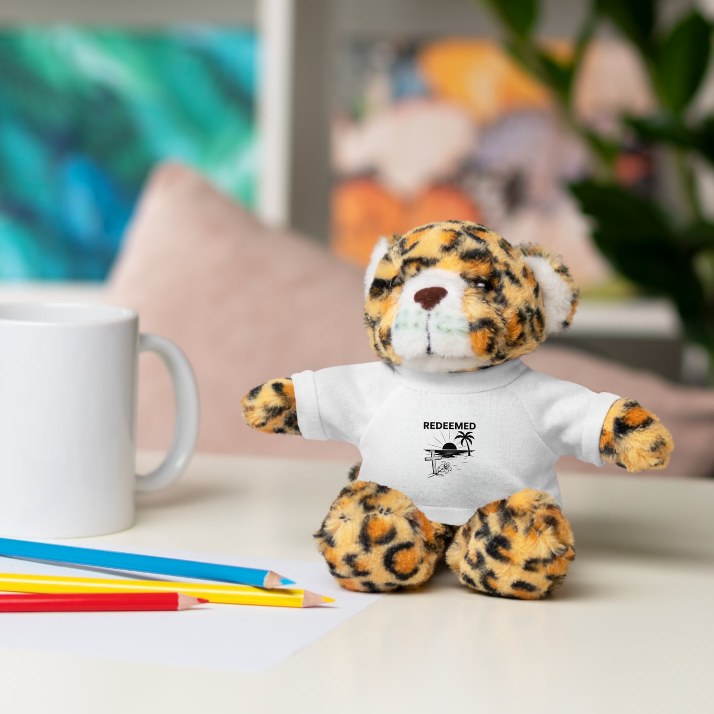 Leopard print teddy bear wearing a white shirt with text, sitting on a desk with stationery items.