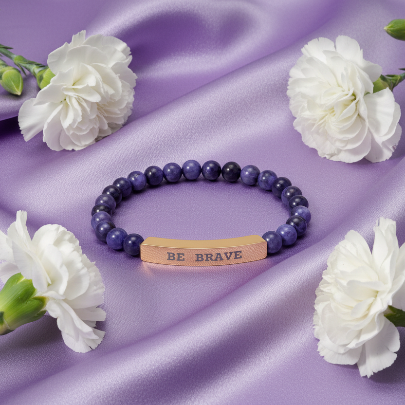 Natural Stone dark blue beaded stretch bracelet with 'BE BRAVE' bar engraved on a purple fabric background with white carnations.