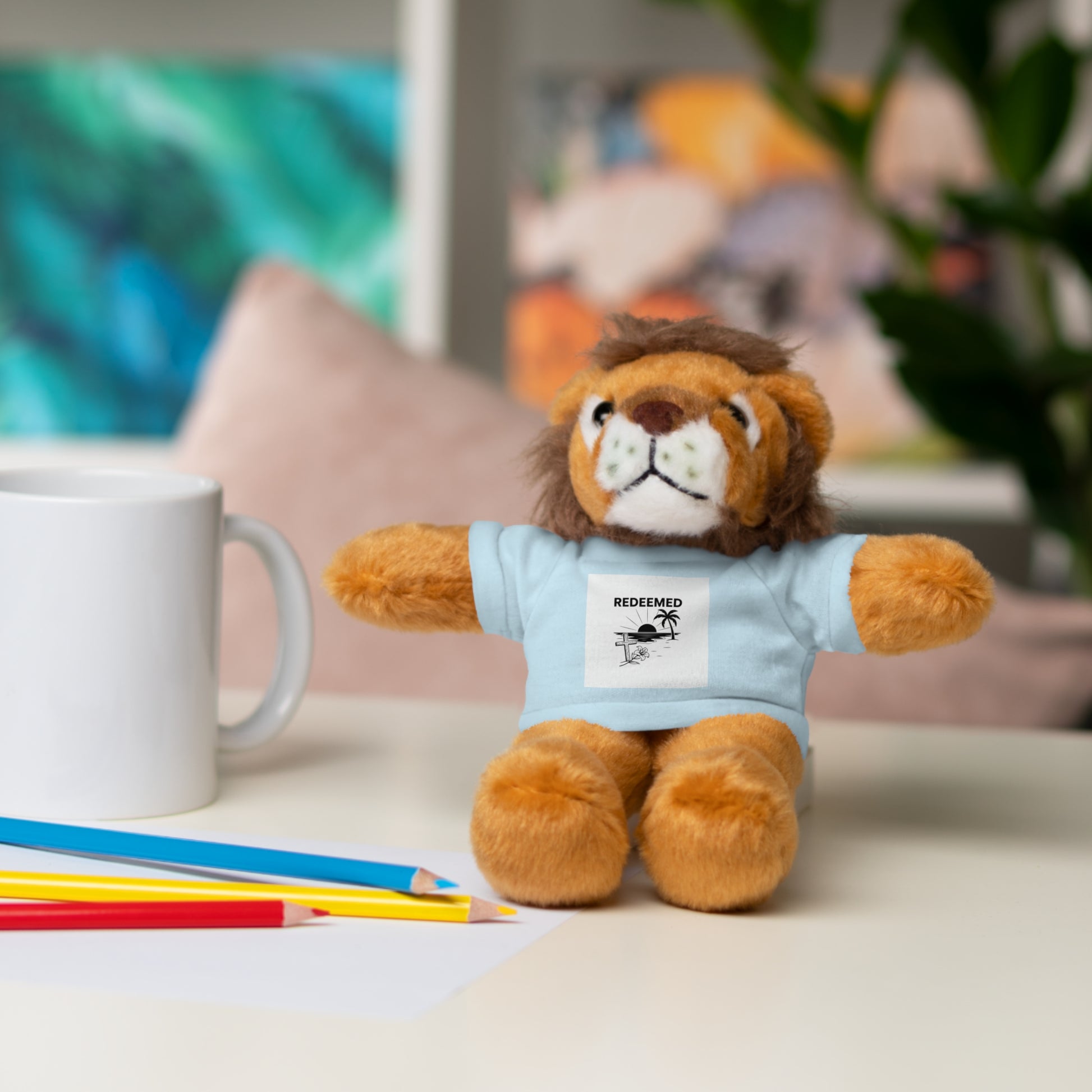Plush lion toy wearing a blue shirt with a graphic on a table with stationery items.