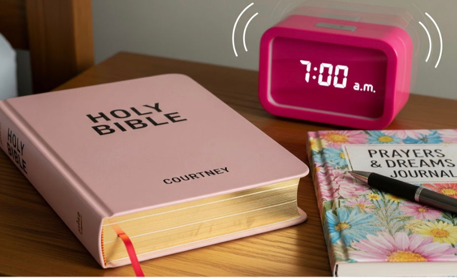 Pink Bible with 'HOLY BIBLE' and 'COURTNEY', pink alarm clock showing 7:00 a.m., and floral journal on a wooden surface.